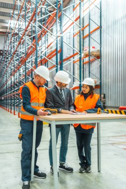 Depo çalışanları ve yöneticileri güvenlik kaskı takıp barkod tarayıcısı ve masa üzerindeki belgeleri kullanarak lojistik operasyonlarını tartışıyorlar.