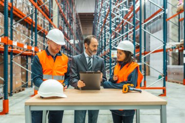 Depo çalışanları ve yönetici büyük bir dağıtım merkezinde bir pano kullanarak lojistiği tartışıyorlar.