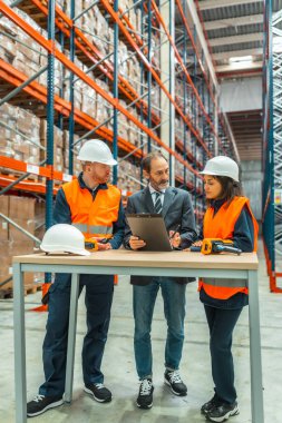 Depo çalışanları ve yönetici büyük bir dağıtım merkezinde bir pano kullanarak lojistiği tartışıyorlar.