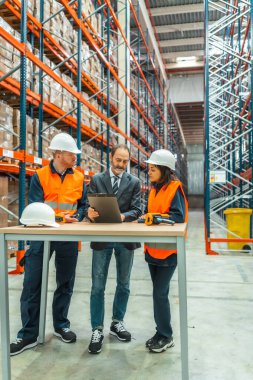 Güvenlik yeleği ve kask giyen depo çalışanlarına envanter durumunu gösteren bir pano var.