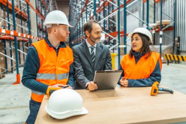 Depo çalışanları ve yönetici büyük bir dağıtım merkezinde bir pano kullanarak lojistiği tartışıyorlar.