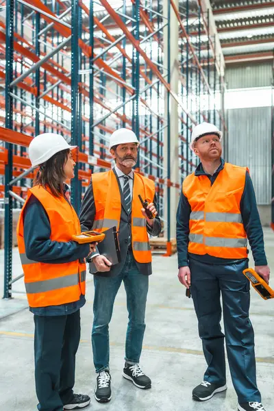 Depo çalışanları ve yöneticileri güvenlik yelekleri ve kasklar takıyor, envanter yönetimi ve lojistik işlemleri için telsiz ve barkod tarayıcısı kullanıyorlar.