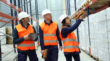 Raflar arasında yürüyen lojistik uzmanları, modern dağıtım merkezinde barkod tarayıcıları kullanarak sistematik olarak envanter kontrolü yapıyorlar, verimli bir şekilde işbirliği yapıyorlar.