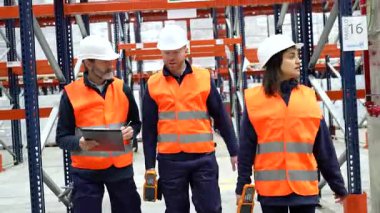 Güvenlik yelekleri ve kask takan depo çalışanları tesise girip lojistiği tartışıyor ve barkod okuyucuları kullanıyorlar.