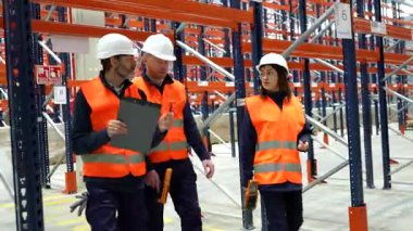 Profesyonel lojistik ekibi depo koridorunda yürüyor, envanter yönetimini analiz ediyor ve operasyon stratejilerini işbirlikçi takım çalışması ile koordine ediyor.