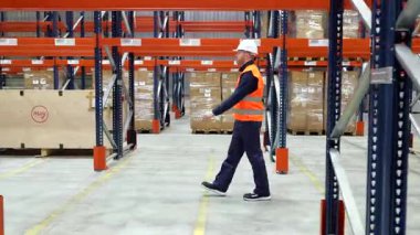 Güvenlik yeleği ve kask giyen lojistik işçisi elinde tarayıcı tutarak depo alanından geçiyor.