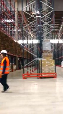 Lojistik işçisi depo koridorlarında yürüyor, envanteri kontrol ediyor ve el bilgisayarı kullanıyor.