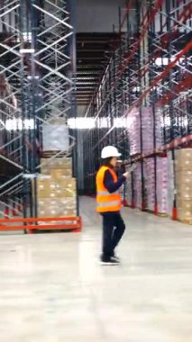 Lojistik operatörü tablet kullanıyor, depo koridorlarında yürüyor, mal ve envanteri kontrol ediyor.