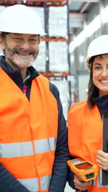 Depoda güvenlik yelekleri ve kasketler takan işçiler gülümseyip dağıtım merkezinde kameraya poz veriyorlar.