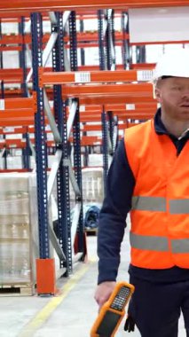 Depo çalışanı çelik yelek ve kasket takıyor depo koridorlarında yürüyor ve envanter kontrolü için el tarayıcısı kullanıyor.