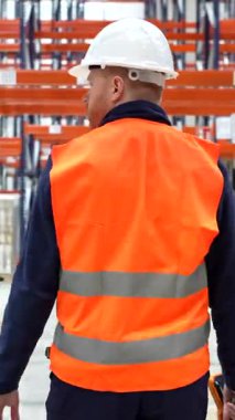 Depo koridorlarında güvenlik ekipmanı taşıyan profesyonel lojistik işçisi, modern dağıtım merkezinde tedarik zinciri operasyonlarını ve envanter yönetimini temsil ediyor.