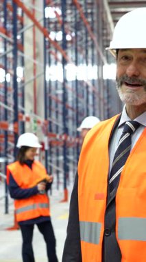 Güvenlik kaskı ve yelek giyen üst düzey yönetici telefonda konuşuyor ve depoda, lojistik ve dağıtım merkezinde gülümsüyor.