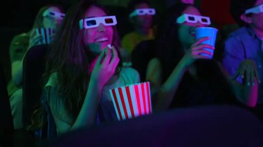 Üç boyutlu gözlük takan çok kültürlü arkadaşlar patlamış mısır paylaşıyorlar, karanlık sinemada film gösterimi sırasında heyecanla sinemalara dalıyorlar.