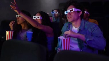 Farklı arkadaşlar 3D gözlük takıyor, sinemada film izlerken uzanıp patlamış mısır yiyorlar.