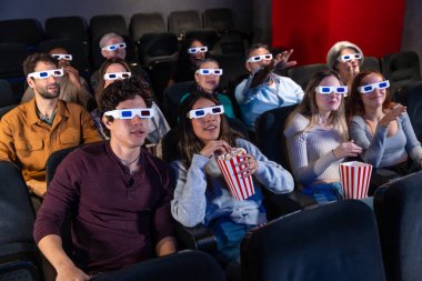 Farklı yaşlarda insanlar 3D gözlük takıyor, patlamış mısır yiyor ve sinemada film seyrediyor, birlikte eğleniyorlar.
