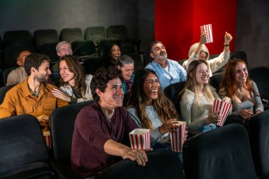 Farklı yaş ve etnik kökenlere sahip insanlar sinemada film izlerken, patlamış mısır yerken gülüyor ve eğleniyorlar.