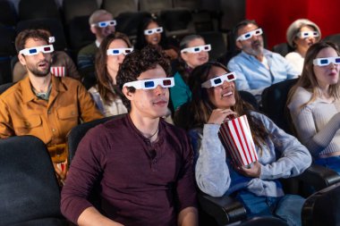 Farklı yaşlarda, 3D gözlüklü insanlar sinemada film izliyor, patlamış mısır yiyor ve gösterinin tadını çıkarıyorlar.