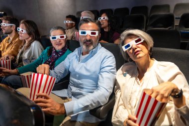Farklı yaş ve etnik kökenlere sahip insanlar 3D gözlük takıyor ve sinemada film izlerken patlamış mısır yiyorlar.