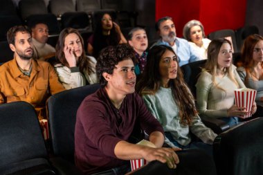 Çeşitli yaş ve etnik kökenlere sahip izleyiciler karanlık bir sinemada heyecan verici bir film gösterimi deneyimlediler, gerilim ve heyecan anlarını paylaştılar.