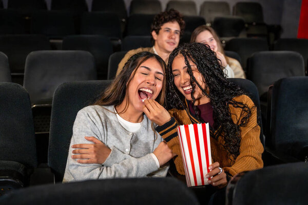 Две молодые женщины наслаждаются фильмом в кинотеатре, смеются и делятся попкорном, с другими людьми на заднем плане также смотрят фильм
