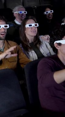 3D gözlük takan insanlar sinemada bir filmden zevk alıyorlar, coşkuyla tepki veriyorlar ve patlamış mısır yiyorlar.