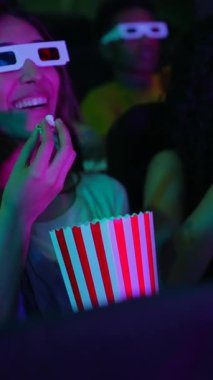 Genç bir kadın, diğer izleyicilerle çevrili bir sinemada 3 boyutlu film izlerken patlamış mısırdan zevk alıyor.