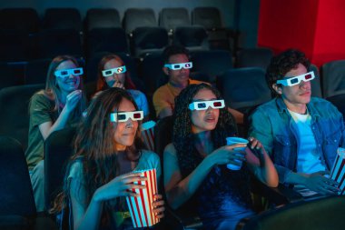3 boyutlu gözlük takan bir grup genç sinemada eğleniyor, patlamış mısır yiyor ve gazoz içiyor.