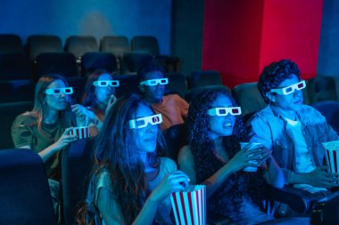 Sinemada 3D film izleyen, 3D gözlük takan ve patlamış mısır yiyen çok ırklı gençler.