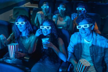 Üç boyutlu gözlük takan ve sinemada patlamış mısır yiyen bir grup genç.