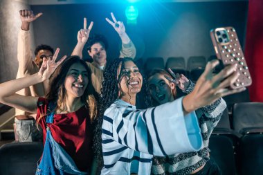 Neşeli gençler sinemada film izlerken akıllı telefonlarıyla selfie çekiyorlar arkadaşlarıyla eğleniyorlar.