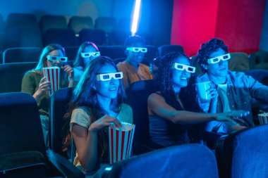 3 boyutlu gözlük takan gençler sinemada film seyrediyor, patlamış mısır yiyor ve gazoz içiyor.