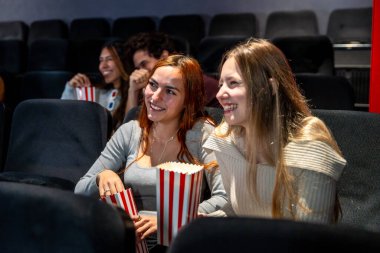 İki genç kadın, sıcak bir sinemada birlikte film izlerken patlamış mısır yiyip gülümsüyor, kahkahalarını paylaşıyor ve eğlenceli bir akşam geçiriyor.
