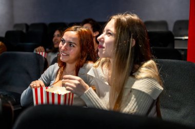Sıcak bir sinemada birlikte film izlerken gülümseyen ve patlamış mısırdan keyif alan iki genç kadın, kahkahalarını paylaşıyor ve eğlenceli bir akşam geçiriyor.