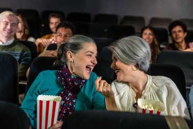 Patlamış mısır paylaşan ve birlikte gülen iki neşeli son sınıf kadını sinemada film izlerken eğleniyorlar.