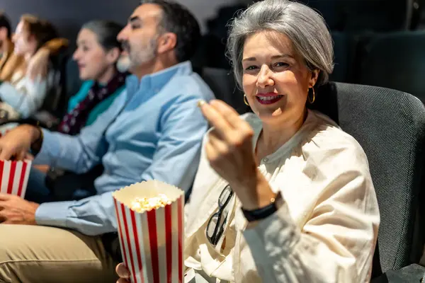 Olgun bir kadın, arkadaşlarıyla sinemada film izlerken gülümseyip patlamış mısır yiyor, boş zamanlarının keyfini çıkarıyor.