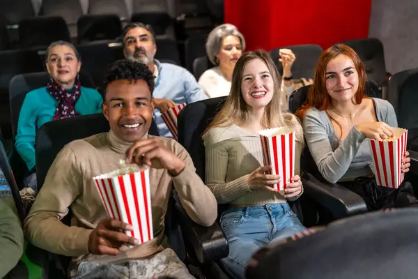 Sinemada eğlenen, kahkahalarını paylaşan ve sıcak bir salonda patlamış mısır yiyen çok ırklı bir arkadaş grubu.