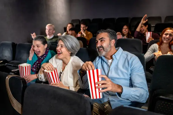 Farklı yaşlarda insanlar sinemada film izlerken gülüyor ve patlamış mısır yiyorlar.