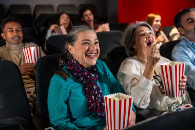 Farklı yaşlarda insanlar sinemada film izlerken gülüyor ve patlamış mısır yiyorlar.