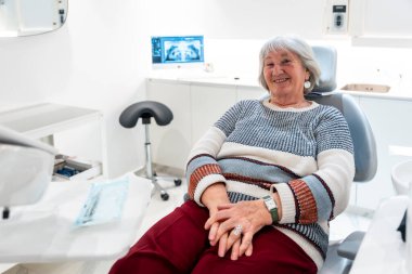 Yaşlı hasta gülümsüyor ve diş muayenesi sırasında rahat hissediyor. Parlak, modern bir klinikte, üst düzey oral bakımı vurguluyor.
