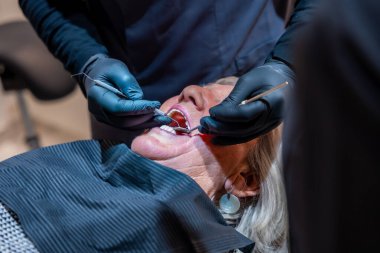 Sağlık kontrolü sırasında dişçi siyah eldiven takmış yaşlı bir kadının dişlerini diş aletleriyle muayene ediyor.