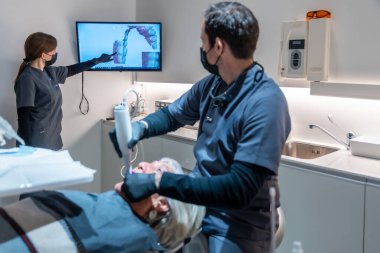 Diş hekimi ve asistanı yenilikçi 3D tarayıcı teknolojisi kullanarak diş beyazlatma prosedürünü gerçekleştiriyor, modern bir klinik ortamında diş bakımını geliştiriyor.