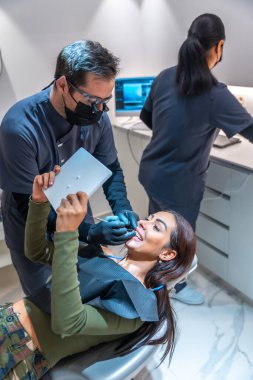 Diş hekimi, arka planda çalışan bir diş hekimi ile gülümseyen bir hastaya tablet üzerinde sonuçlar göstermektedir.