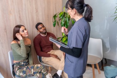 Hemşire tableti tutuyor ve diş kliniğinin bekleme odasında genç çifte tedaviyi anlatıyor.