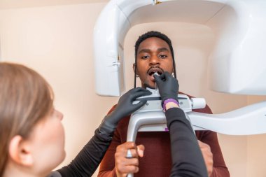 Diş hekimi genç adamın ağzına ısırık bloğu yerleştiriyor, onu modern bir diş kliniğinde diş panoramik radyografisine hazırlıyor.