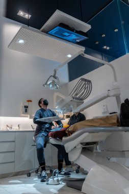 Modern diş kliniğinde hastanın dişlerini inceleyen diş hekimi, gelişmiş teçhizat ve teknolojiyi kapsamlı oral bakım için kullanıyor.