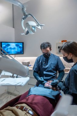 Maske ve eldiven takan dişçi ve asistan modern bir diş kliniğinde bir hastaya diş muayenesi yapıyor.