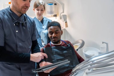 Diş hekimi ve hemşire, dijital tablet üzerinde diş taraması sonuçlarını modern klinikteki hastaya gösteriyor.