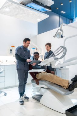 Modern bir diş sağlığı kliniğinde dişçi koltuğunda yatan bir hastaya dijital tablet hakkında bilgi veren dişçi ve diş hemşiresi