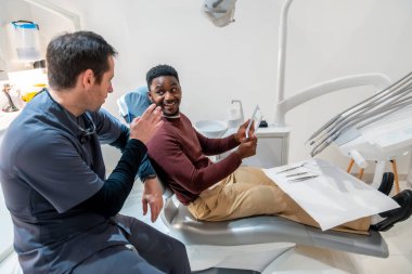 Diş hekimi, dişçi koltuğunda oturan bir hastaya diş röntgenini açıklıyor. Modern bir diş kliniğinde tedavi planını tartışıyor.