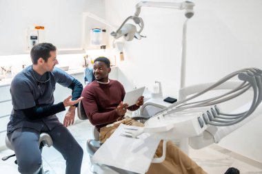 Dişçi, modern diş kliniğinde dijital tablet kullanan hastayla tedavi planını tartışıyor, ileri teknoloji ve hasta bakımını sergiliyor